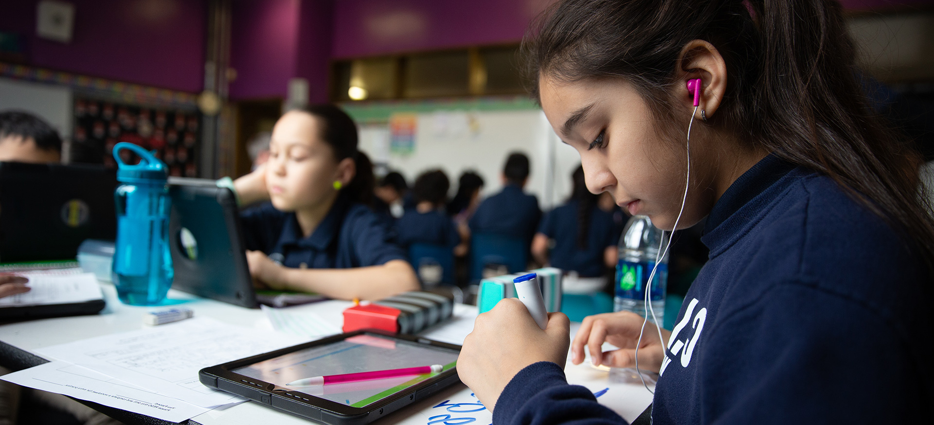 Students working independently at a table in a classroom. One student is wearing headphones and writing with a marker.