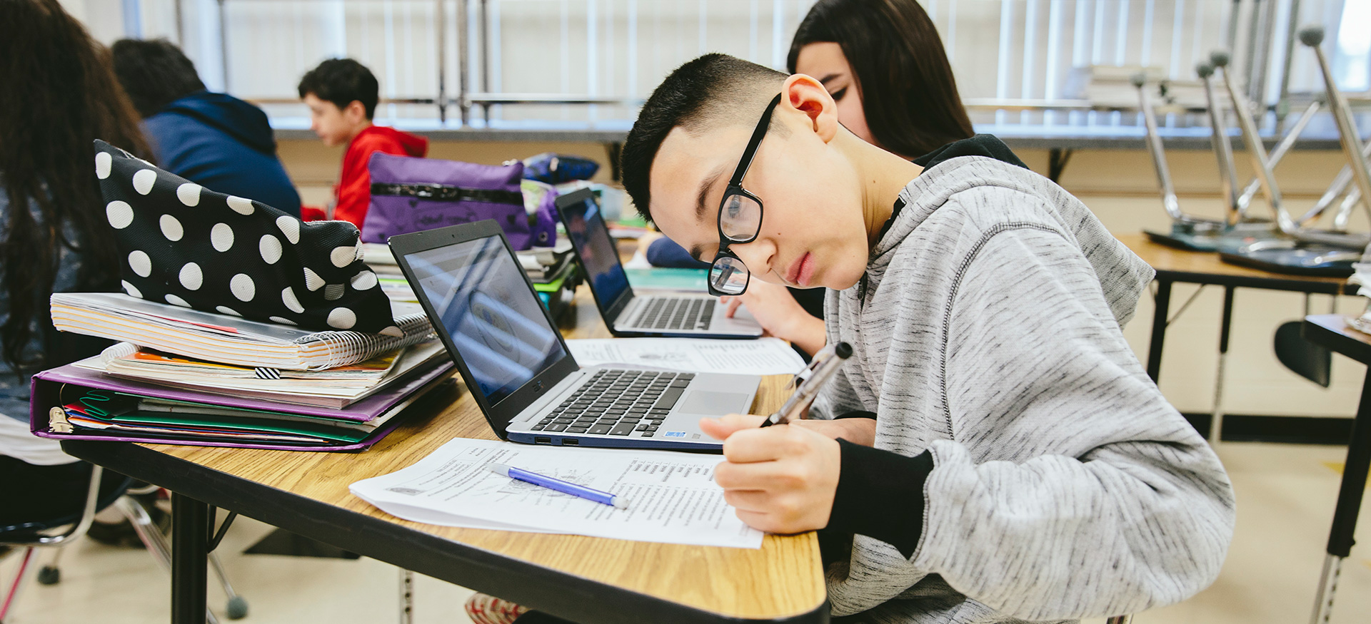 Students working independently at their desks with laptops. One student is leaning over and writing on their paper.