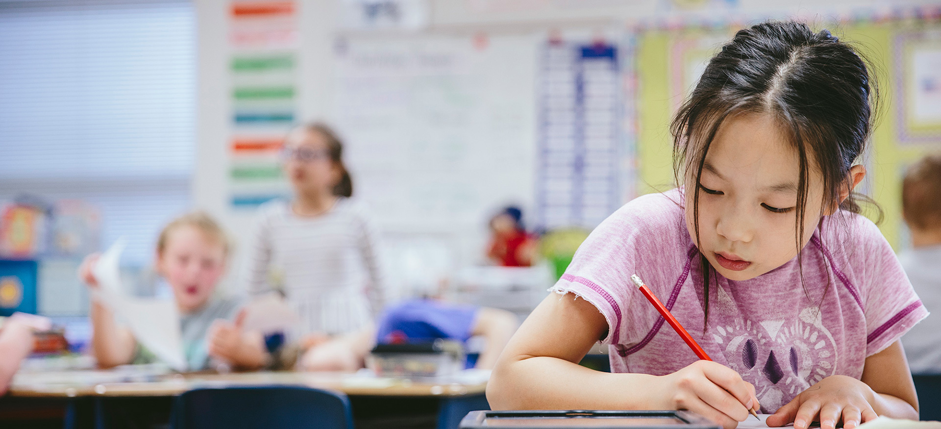 Young student at their desk in a classroom writing with a pencil on paper.