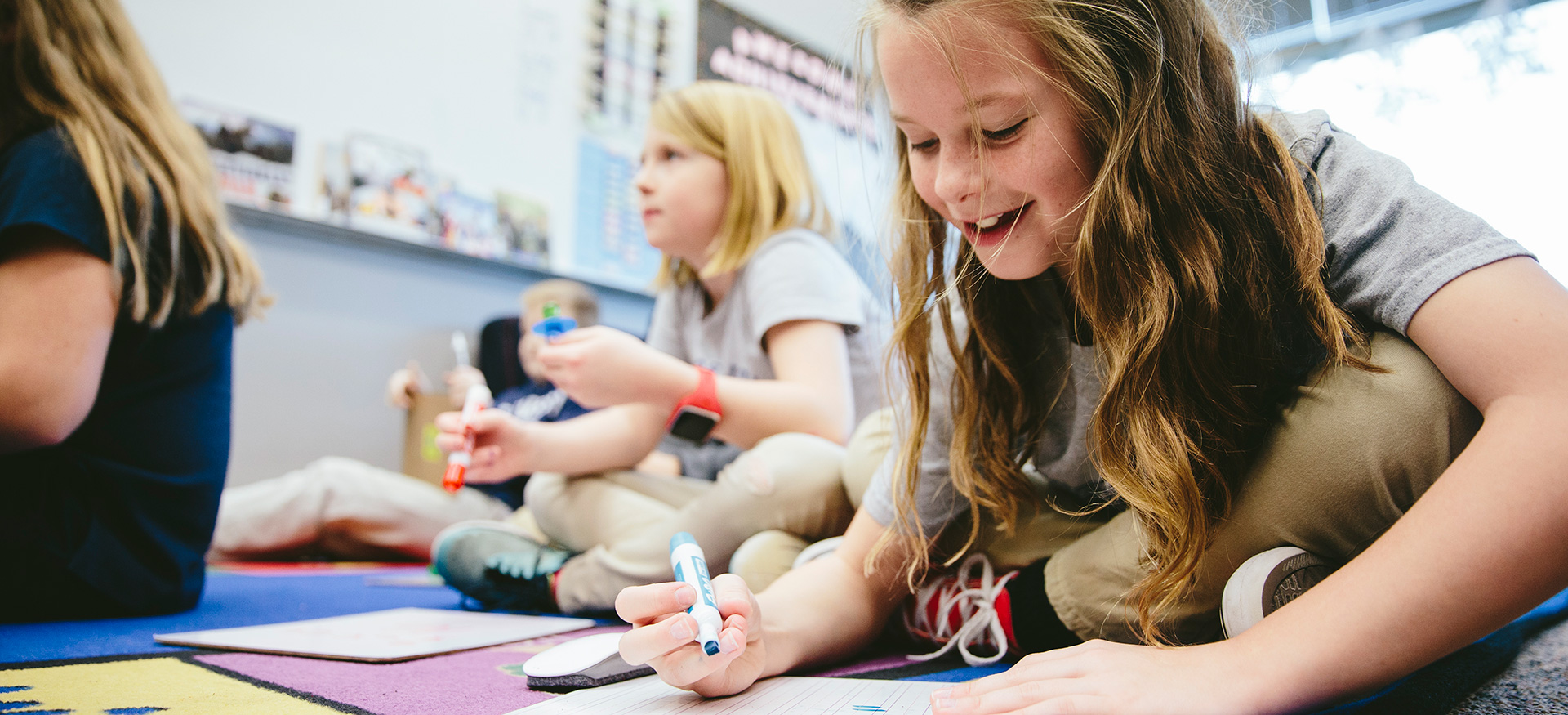 Students sitting on the floor of a classroom working on math problems. One student is looking down and smiling.