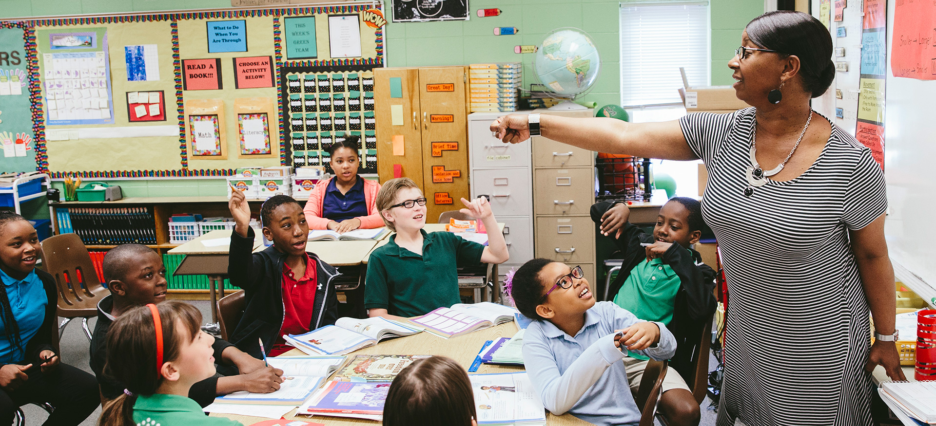 Teacher at the front of a classroom calling on students who are raising their hands.