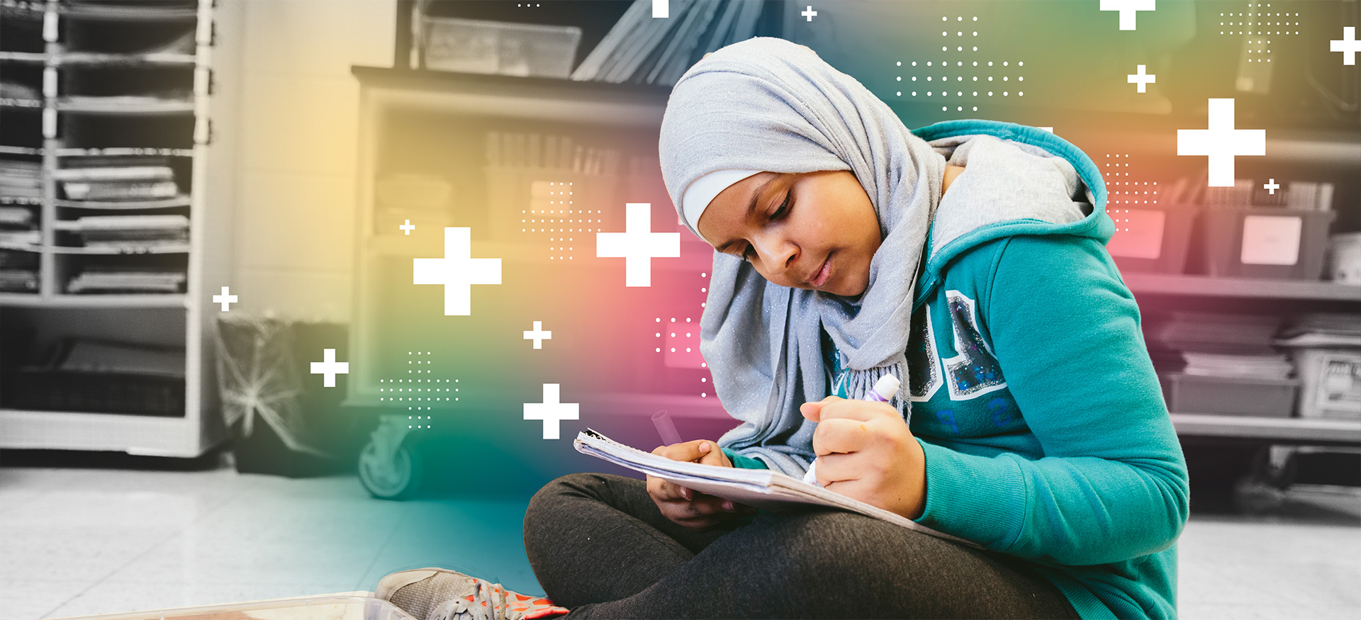 Student sitting crosslegged on a classroom floor writing in a notebook with a marker.