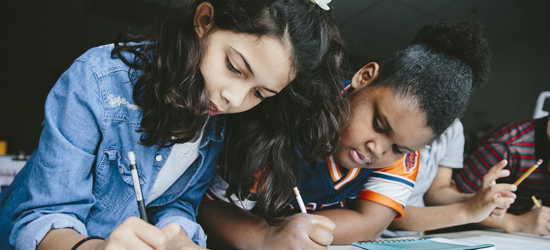 Students at a table working alongside each other. They are writing with pencils on paper.