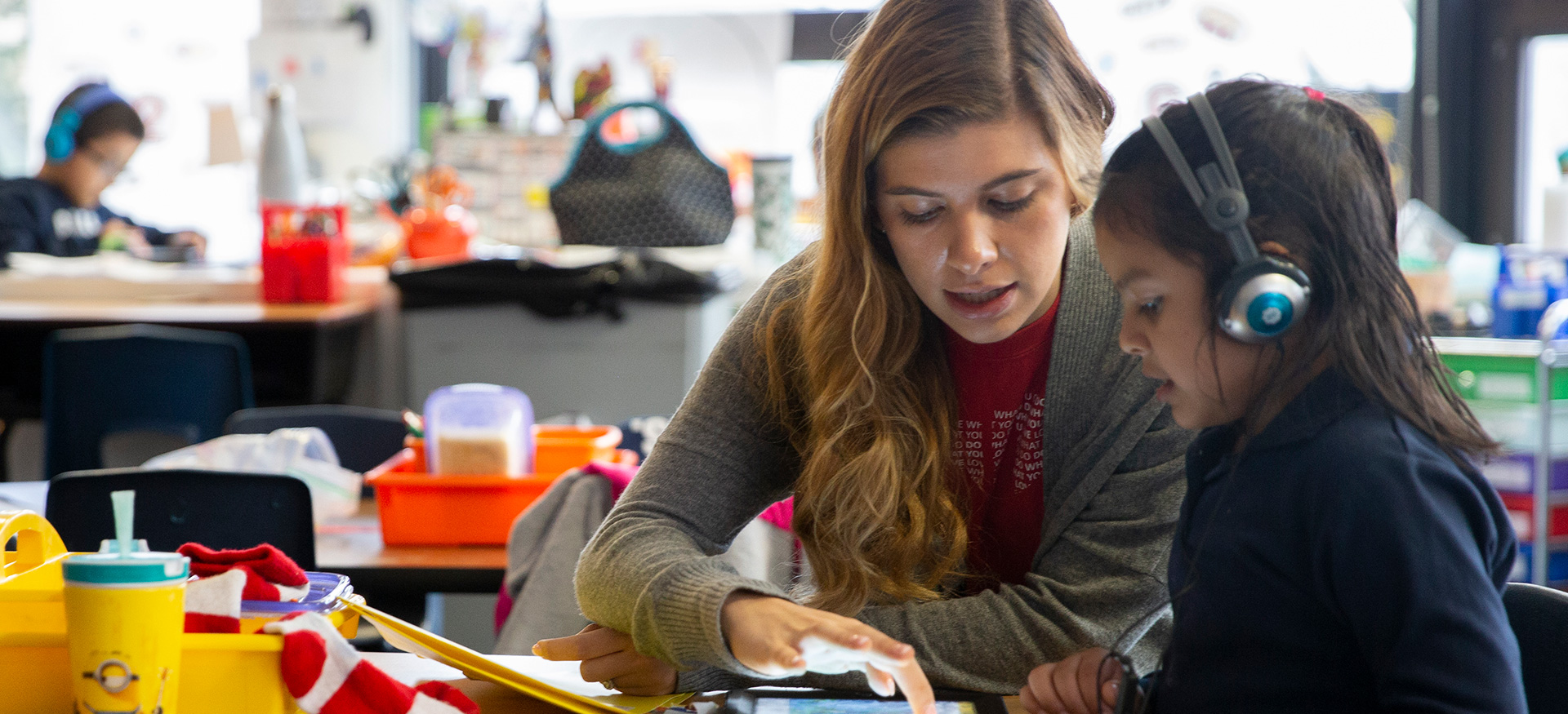 Teacher helping a young student who is wearing headphones and working on a tablet.